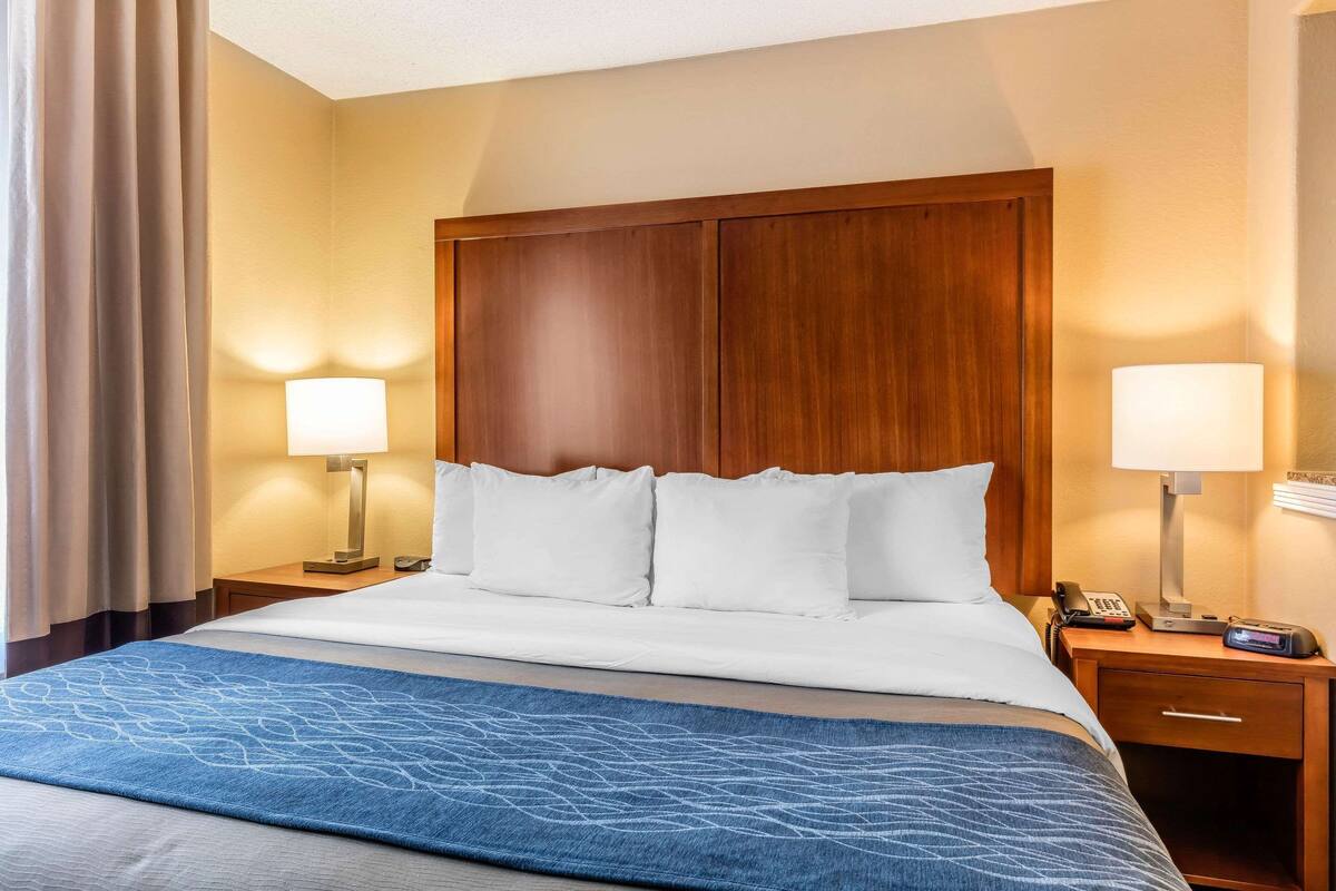 Habitación de hotel con una cama grande, dos lámparas de noche, un cabecero de madera, una mesita de noche con un teléfono y un dispositivo digital, y una manta de color azul con estampado.