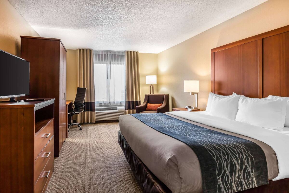 Habitación de hotel con una cama grande, un escritorio con televisor, una silla y una ventana con cortinas.