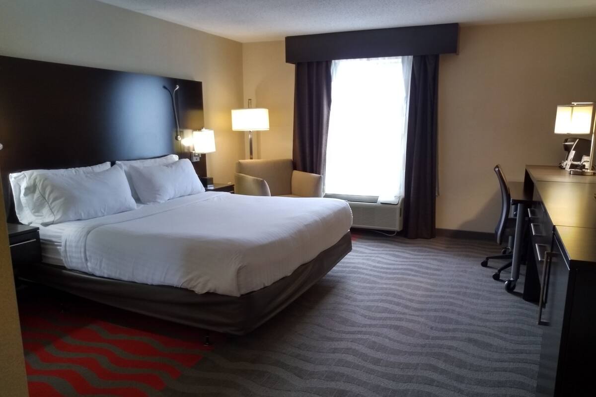 A hotel room with a large bed, a desk, a chair, a lamp, and a window with curtains.