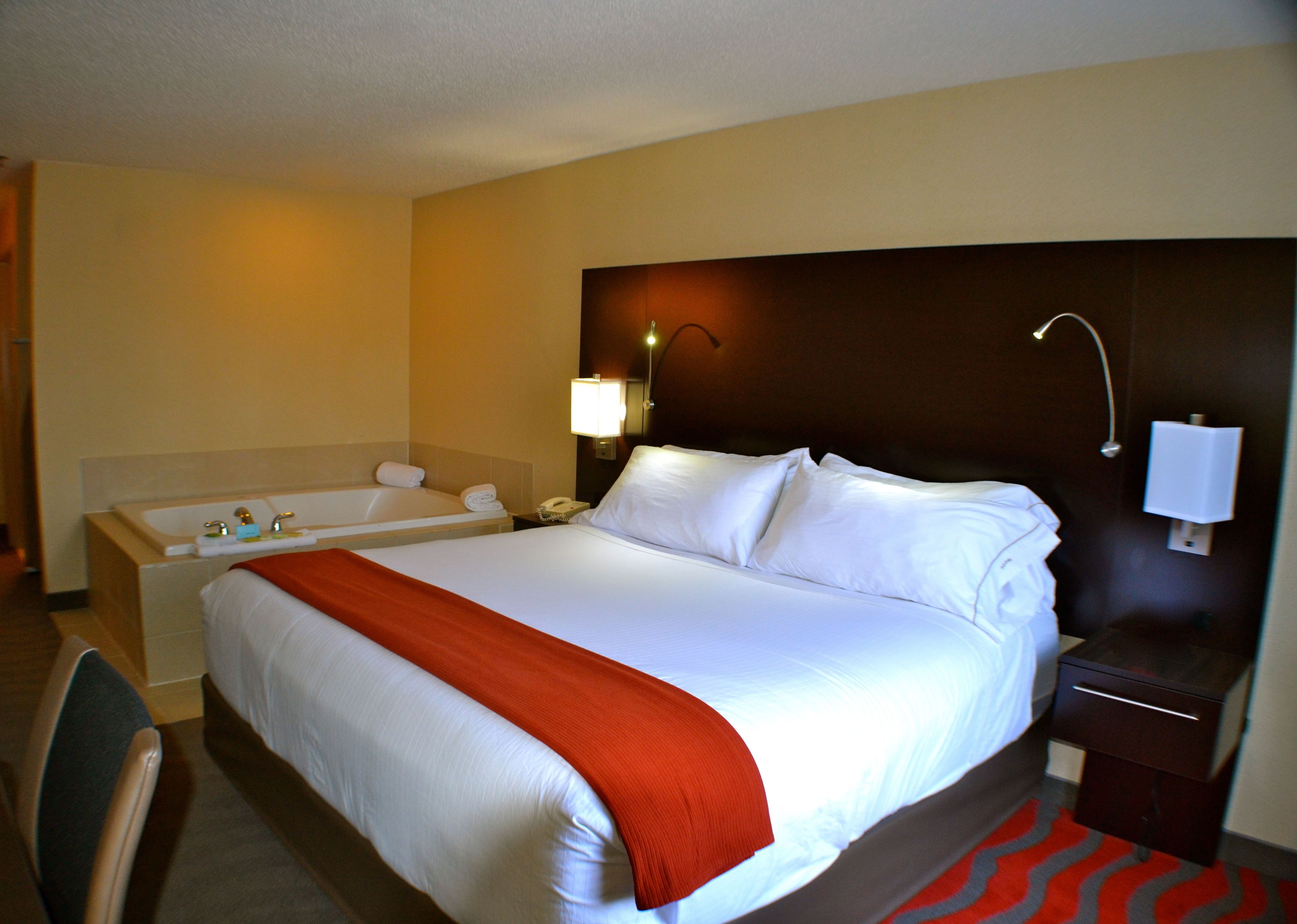 A hotel room with a large bed, a separate bathtub, a bedside table with a lamp, and a red and gray patterned rug.