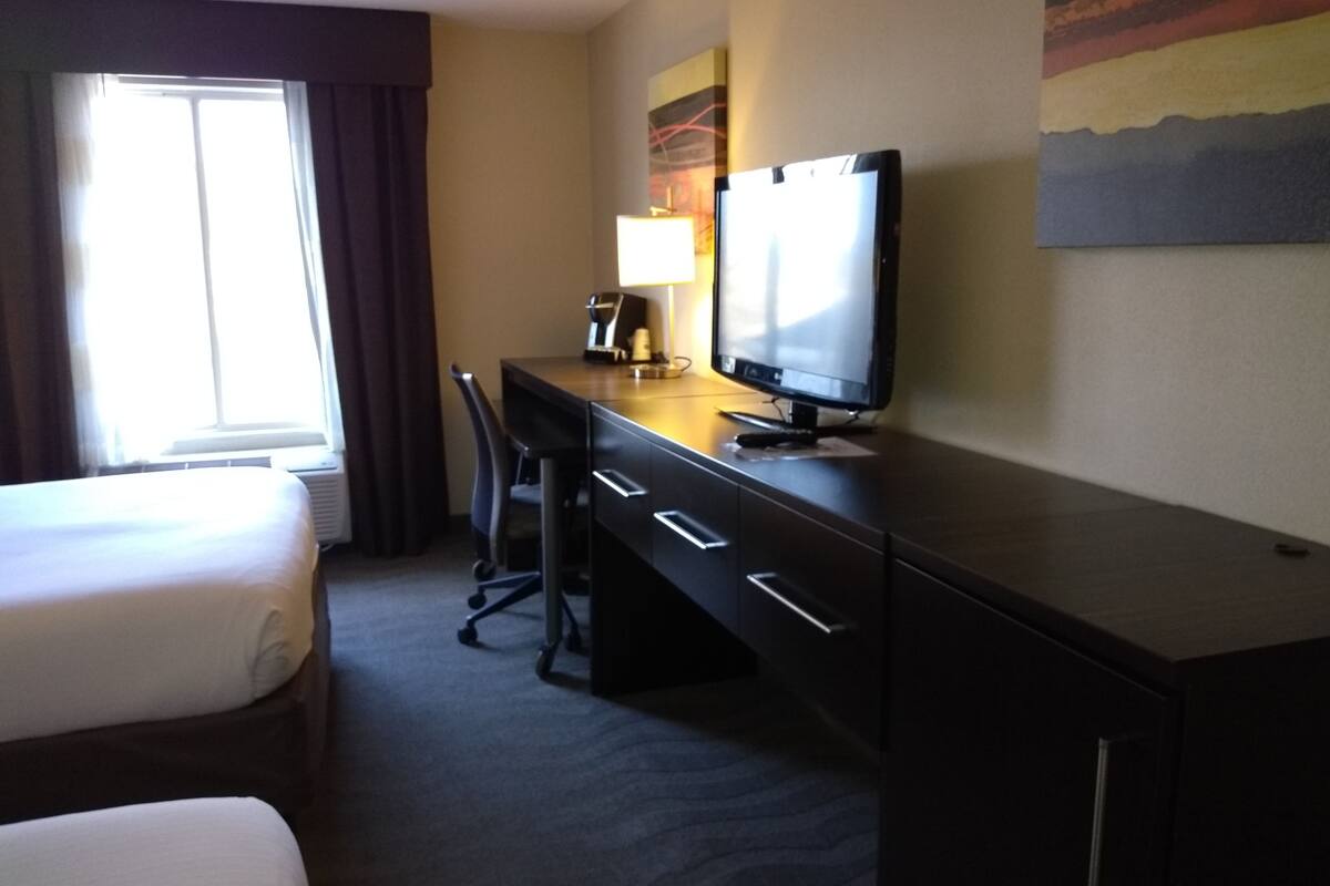 A hotel room with a bed, a desk with a computer, a chair, and a window with curtains.