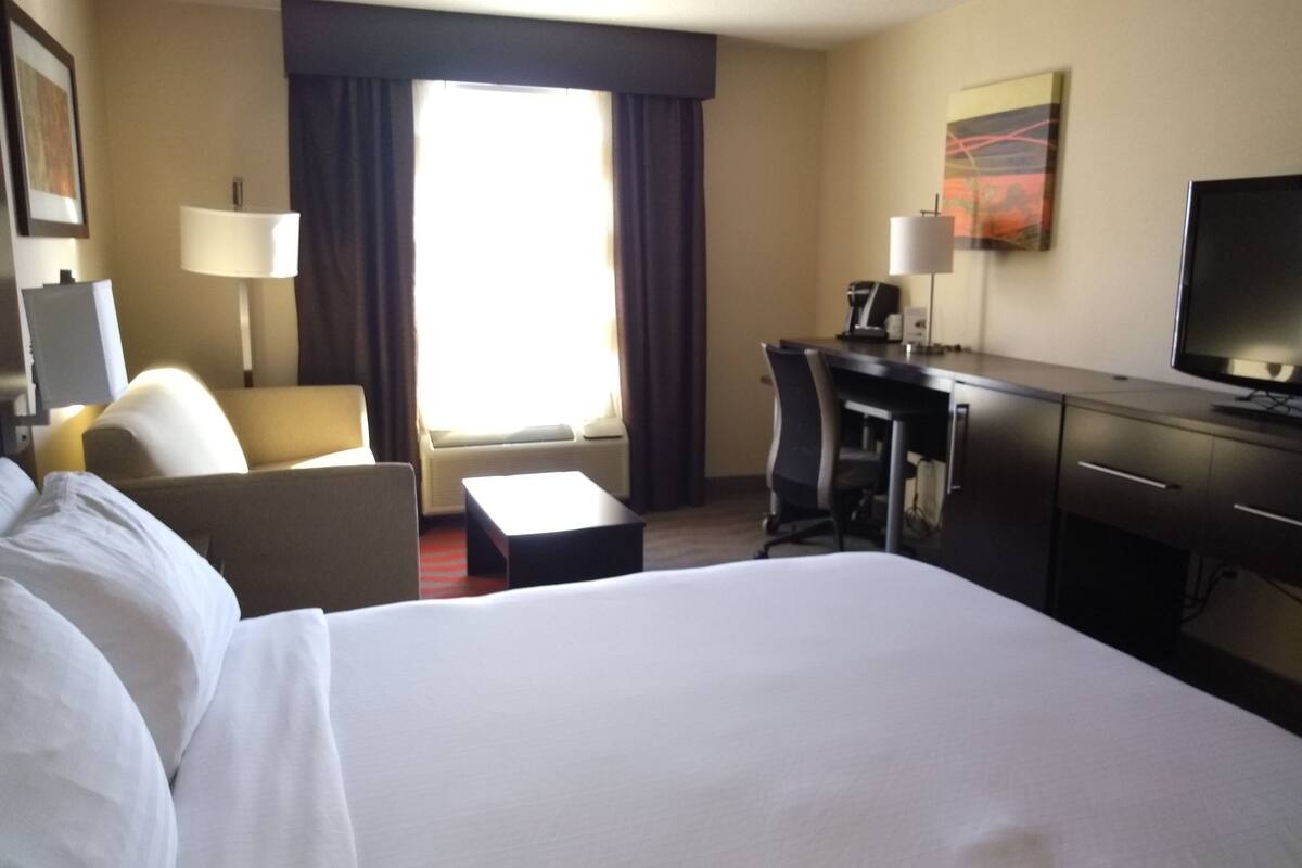 A hotel room with a large bed, a desk, a chair, a TV, and a window with curtains.