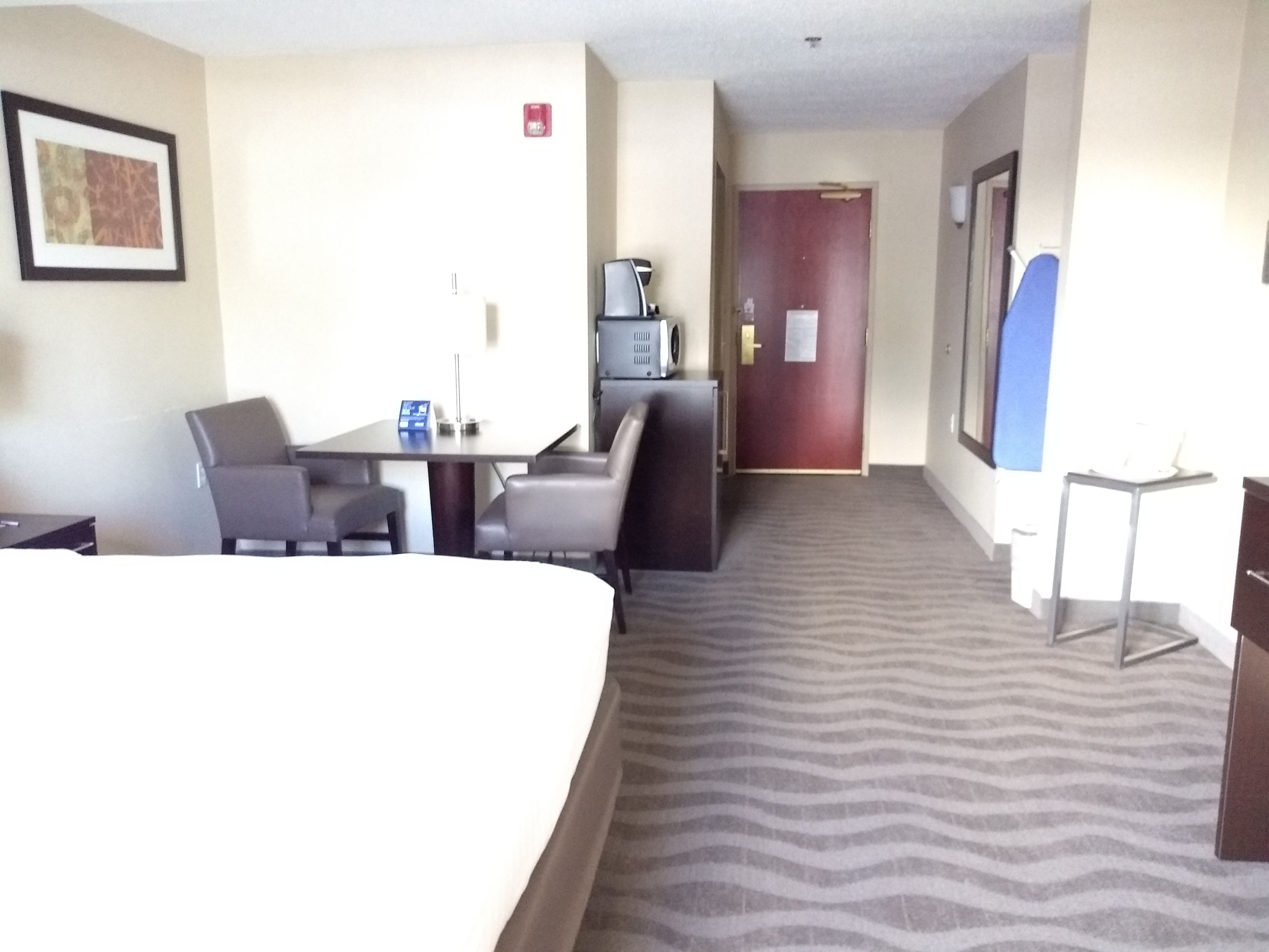 Quarto de hotel com uma cama, uma escrivaninha com cadeira, uma pequena cozinha e uma porta que dá para outro cômodo.