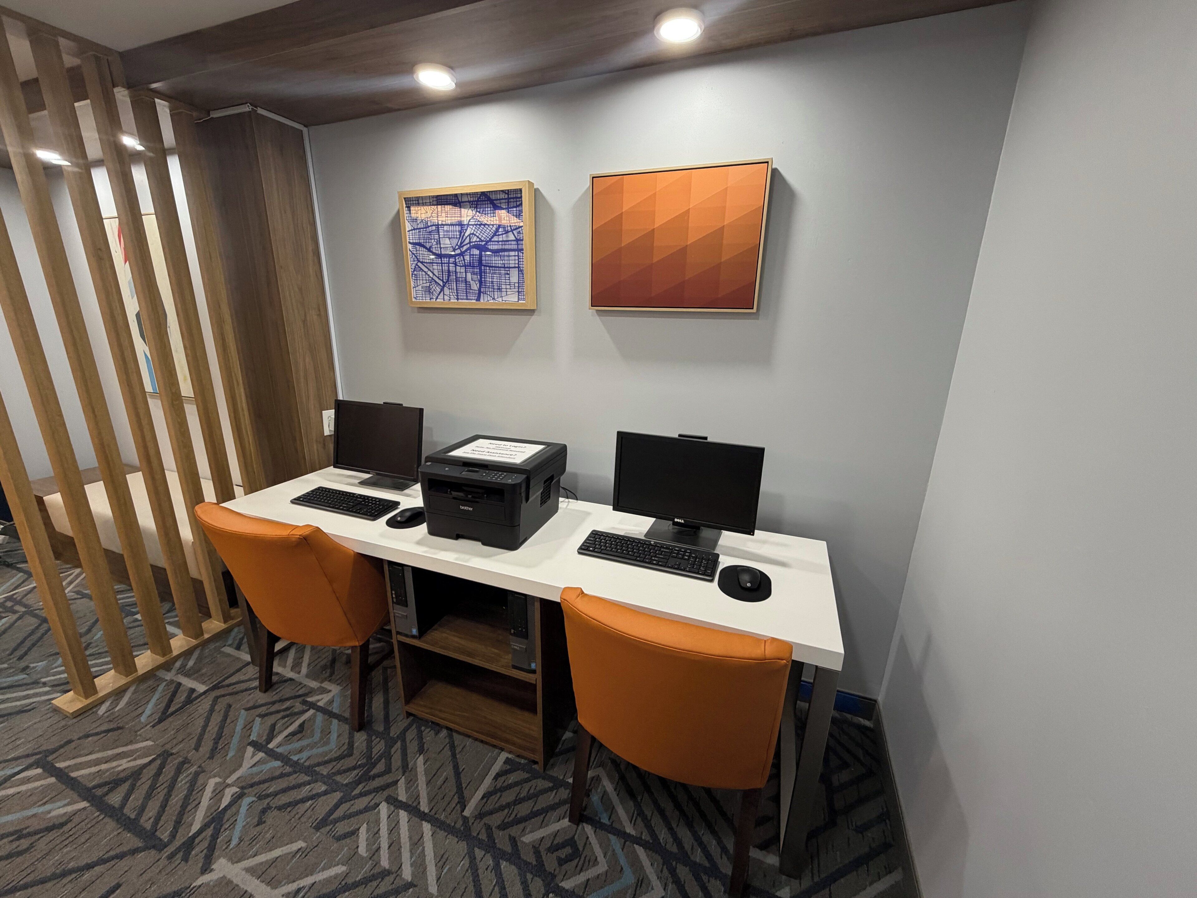 Ein Hotelzimmer mit Schreibtisch, zwei Monitoren, einem Drucker und einer Tastatur. Zwei Stühle und eine dekorative Wandverkleidung sind ebenfalls vorhanden.