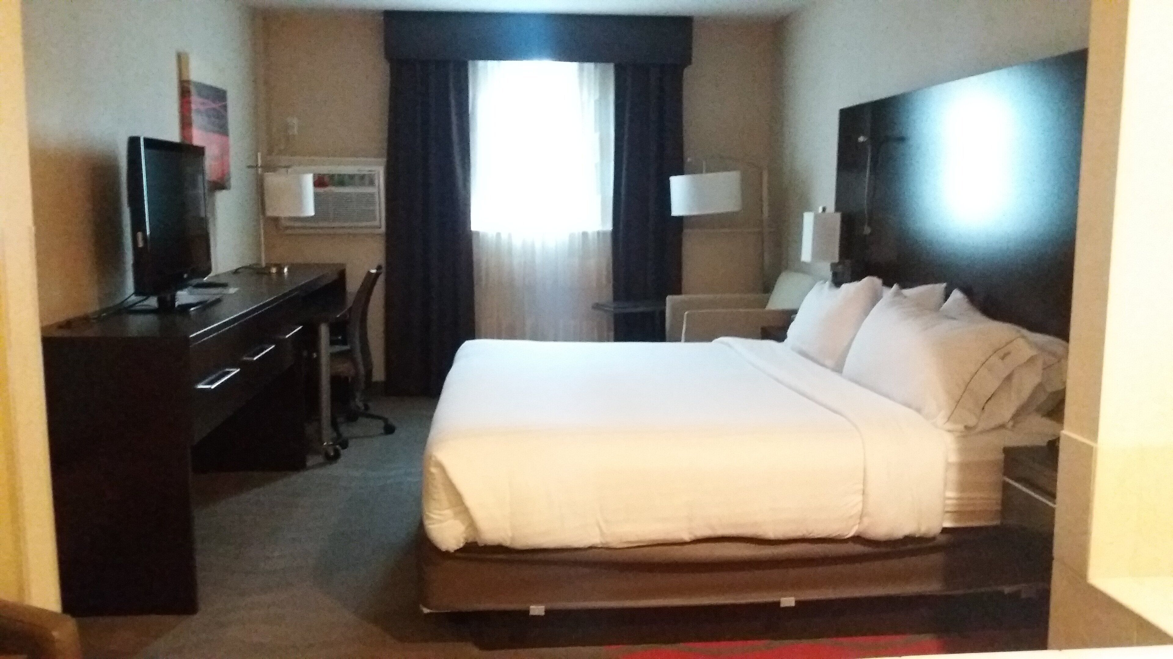 Una habitación de hotel con cama, escritorio, silla, televisor y ventana con cortinas.