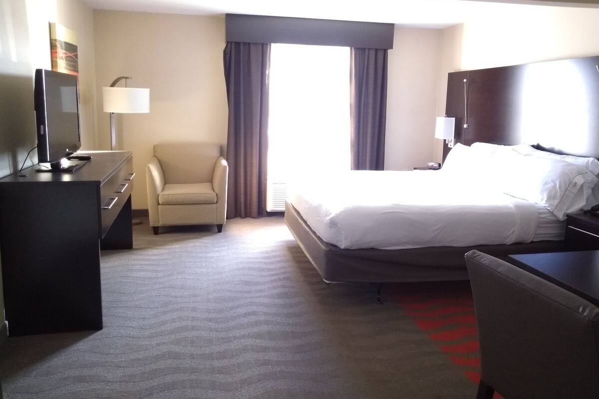 A hotel room with a bed, a desk with a TV, a chair, and a window with curtains.
