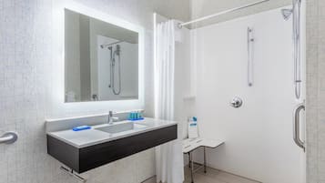A modern bathroom with a large mirror, a sink with a dark countertop, a shower with a glass door, and a small white chair.