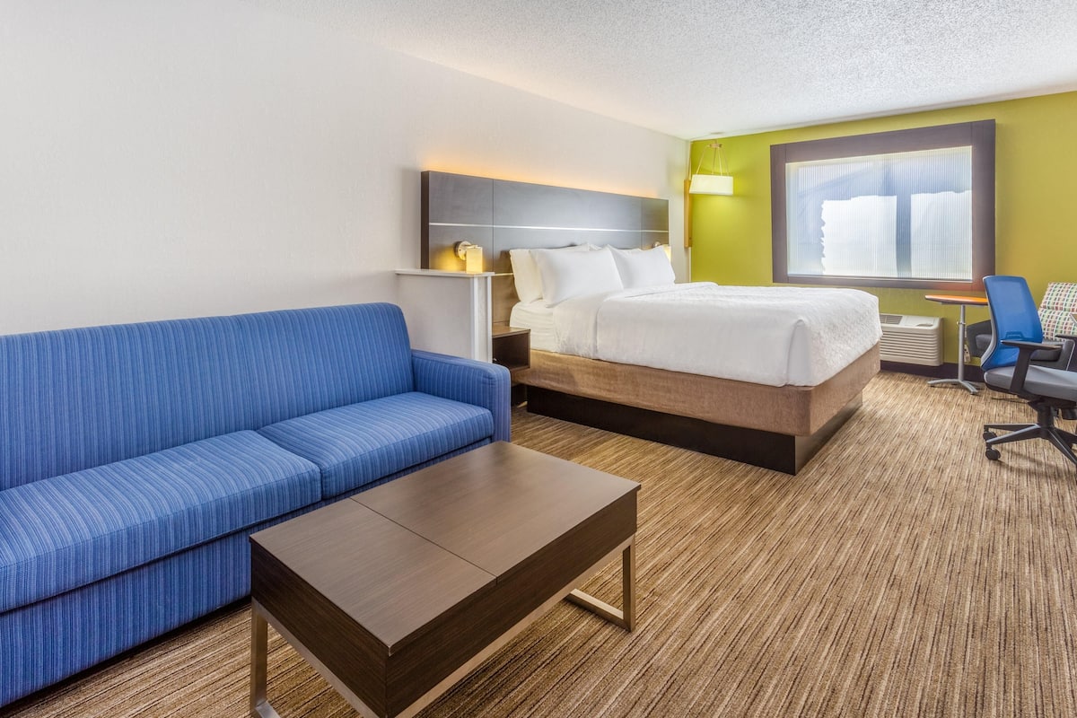 Hotellihuone, jossa on sininen sohva, puinen sohvapöytä, valkoisilla vuodevaatteilla varustettu sänky, yöpöytä lampun kanssa ja ikkuna, jossa on sälekaihtimet.