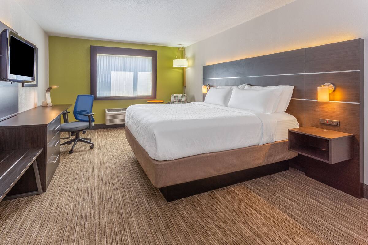 A hotel room with a large bed, a desk with a chair, a television, and a window with blinds.