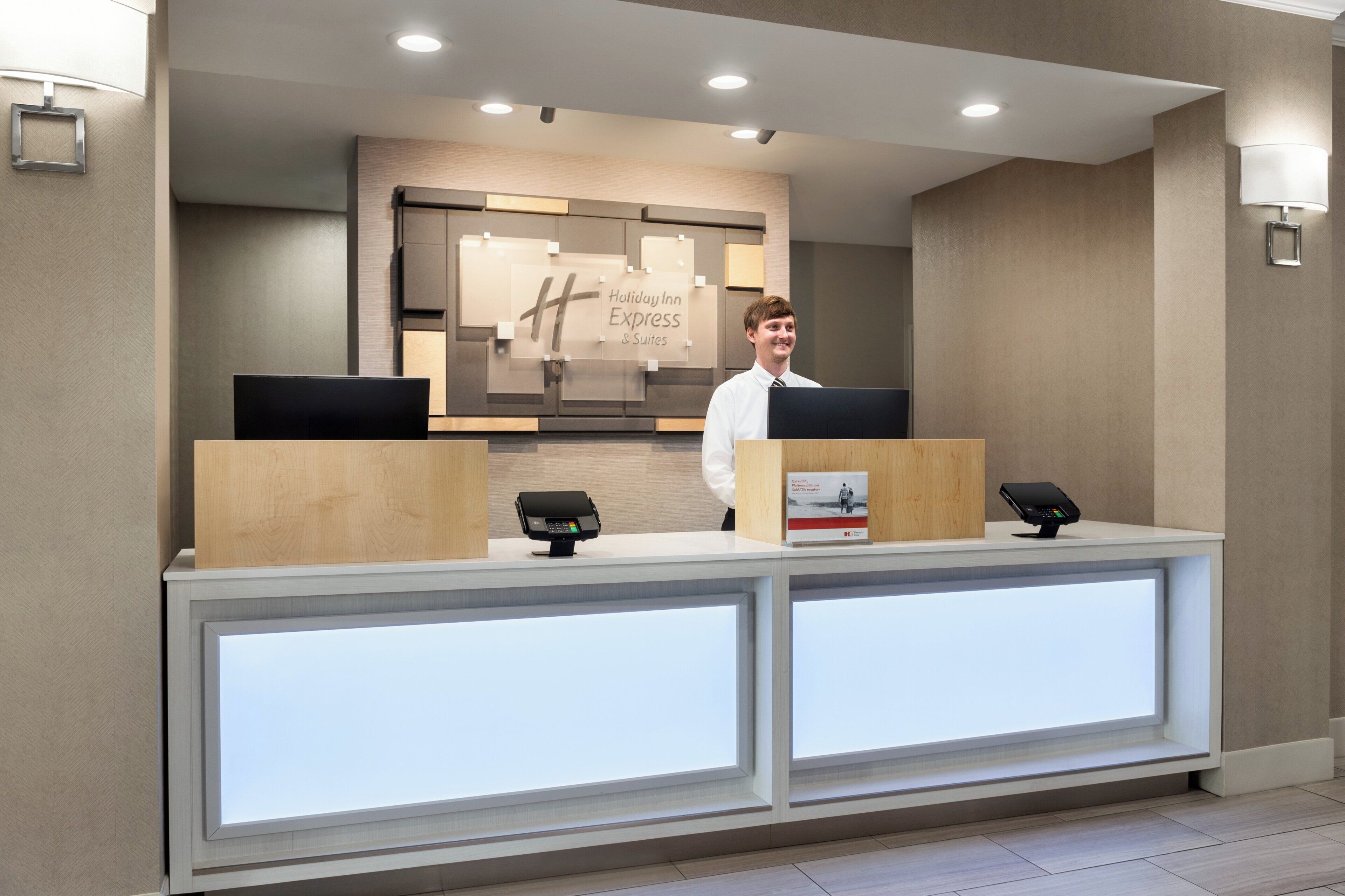 Un mostrador de recepción de hotel con diseño moderno, que incluye un gran letrero de Holiday Inn Express & Suites, dos monitores de computadora y un quiosco digital.