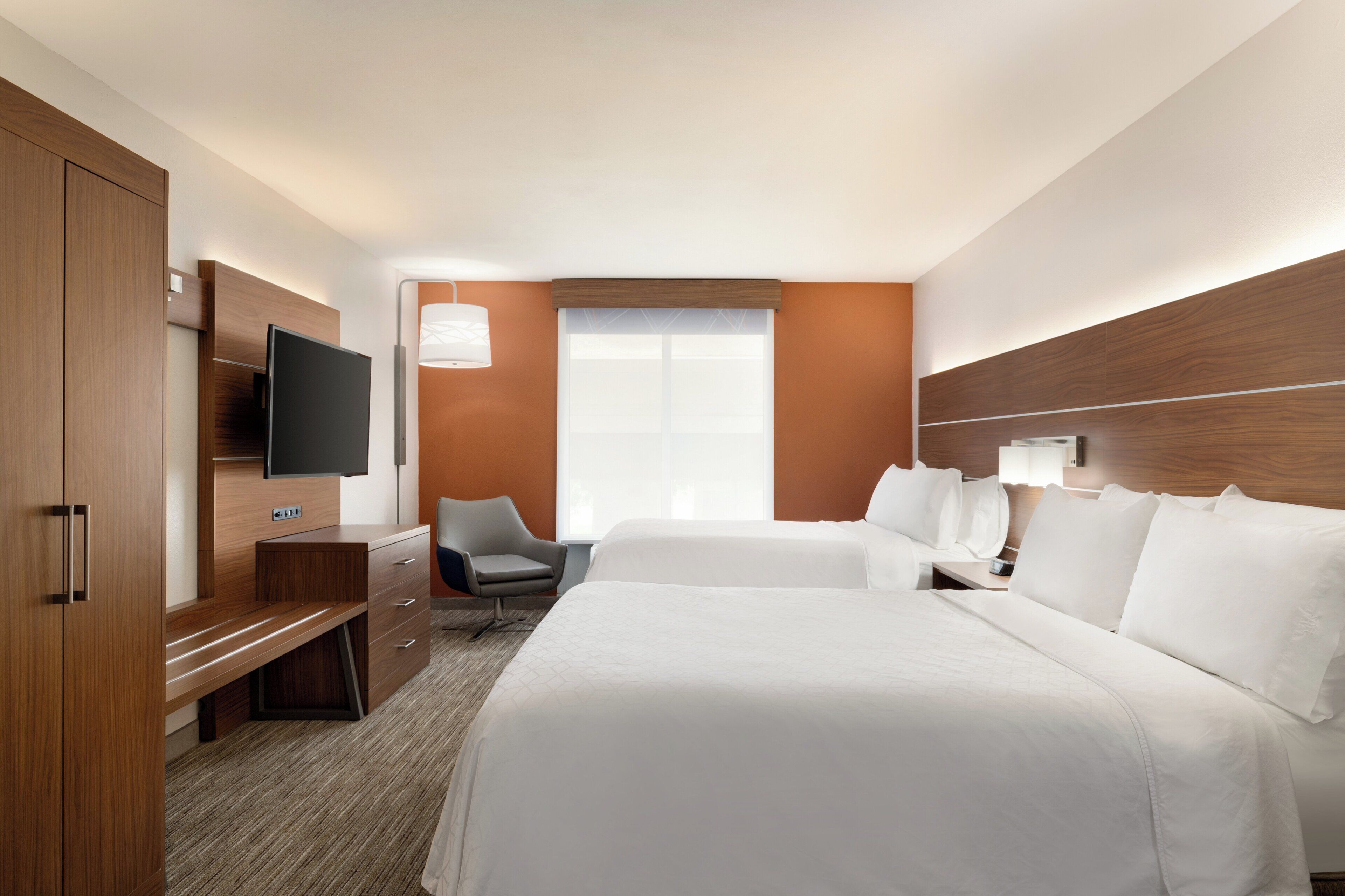 A hotel room with a large bed, a desk with a flat-screen TV, a chair, and a window with blinds.