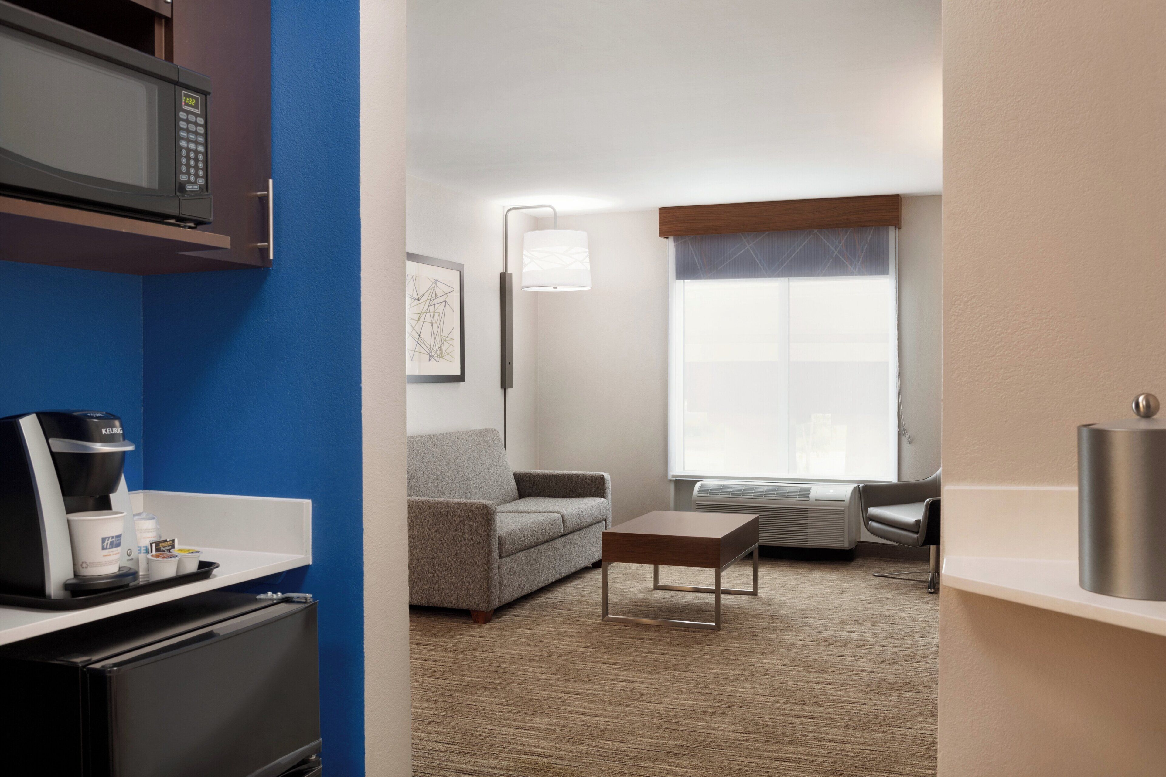 Ein Hotelzimmer mit Küchenzeile, einem grauen Sofa, einer Kaffeemaschine, einem kleinen Tisch und einem großen Fenster mit Jalousien.