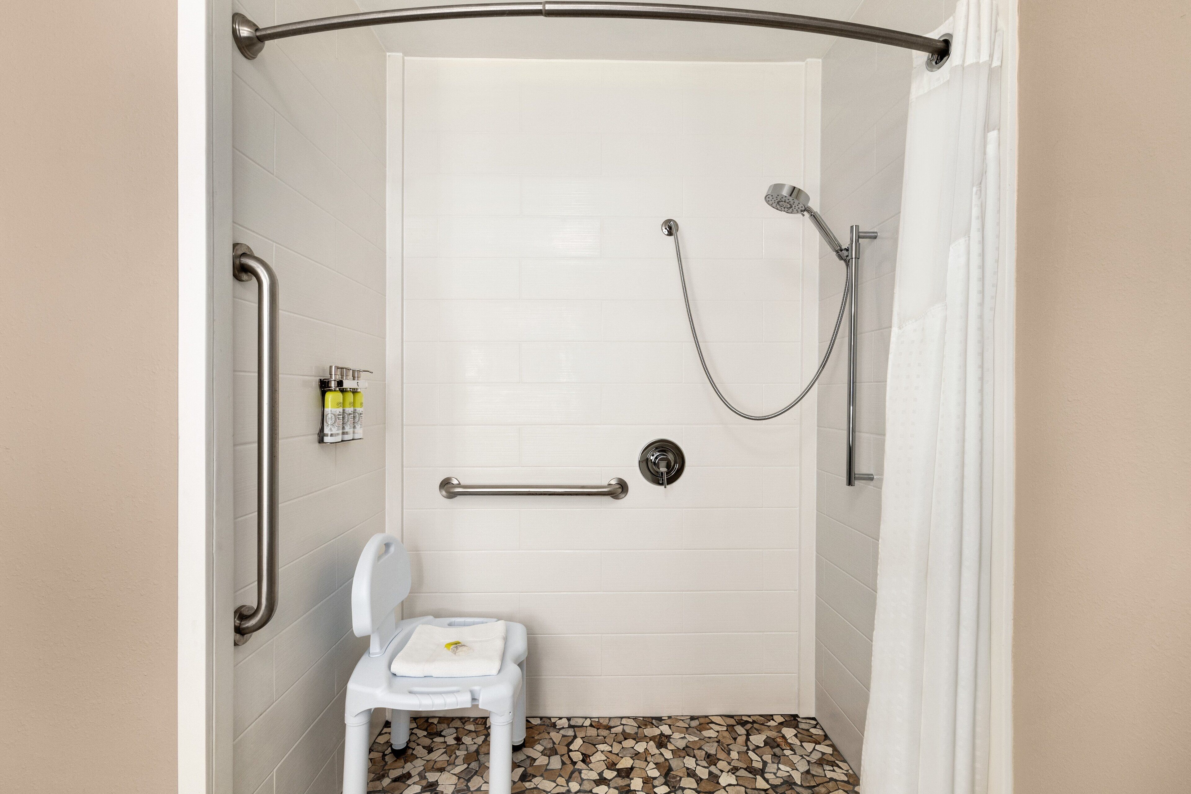 Una cabina de ducha con puerta blanca, barras de apoyo, un cabezal de ducha manual y una pequeña silla blanca con una toalla.