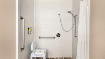 Una cabina de ducha con puerta blanca, barras de apoyo, un cabezal de ducha manual y una pequeña silla blanca con una toalla.