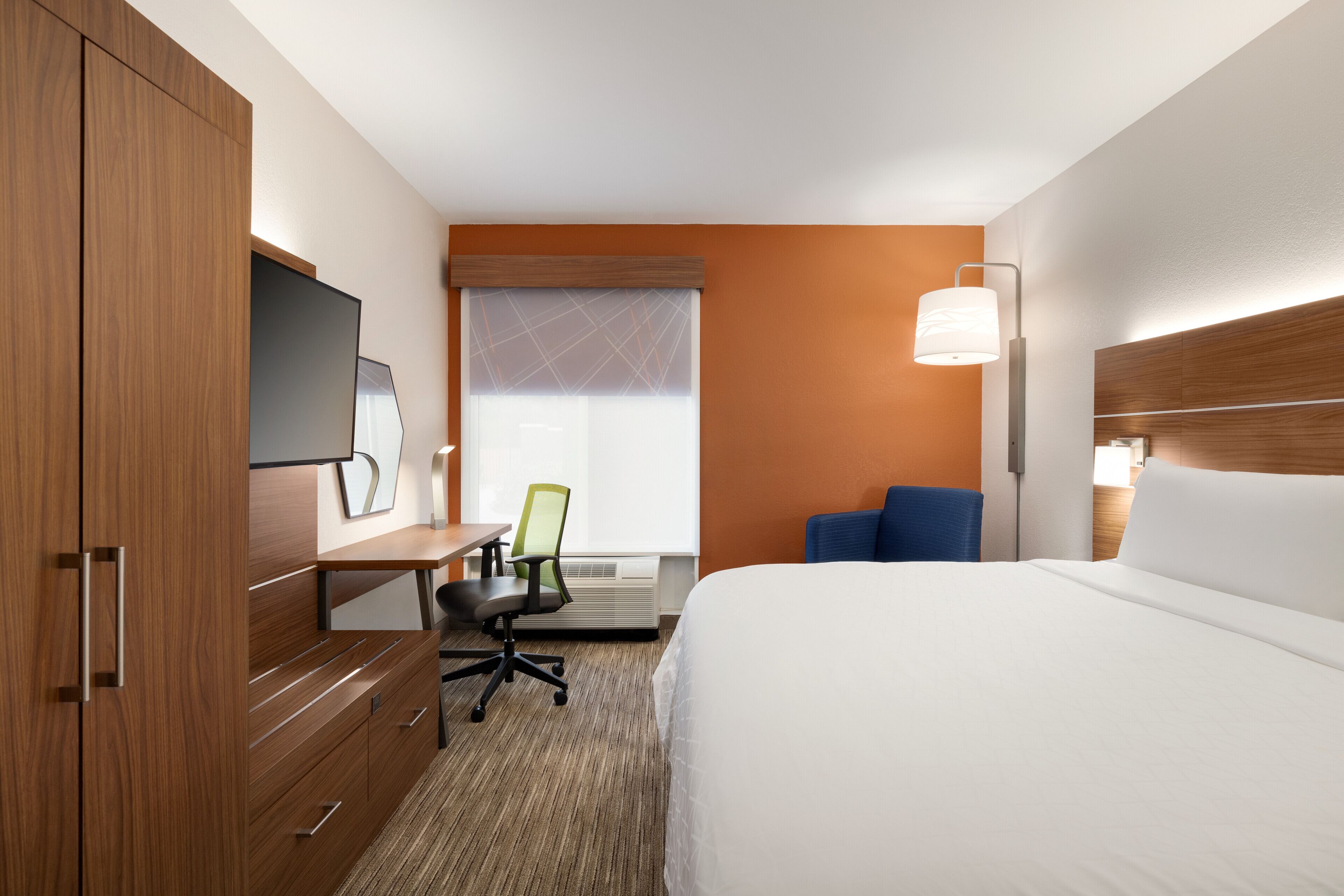 A hotel room with a bed, a desk with a chair, a TV, and a window with blinds.