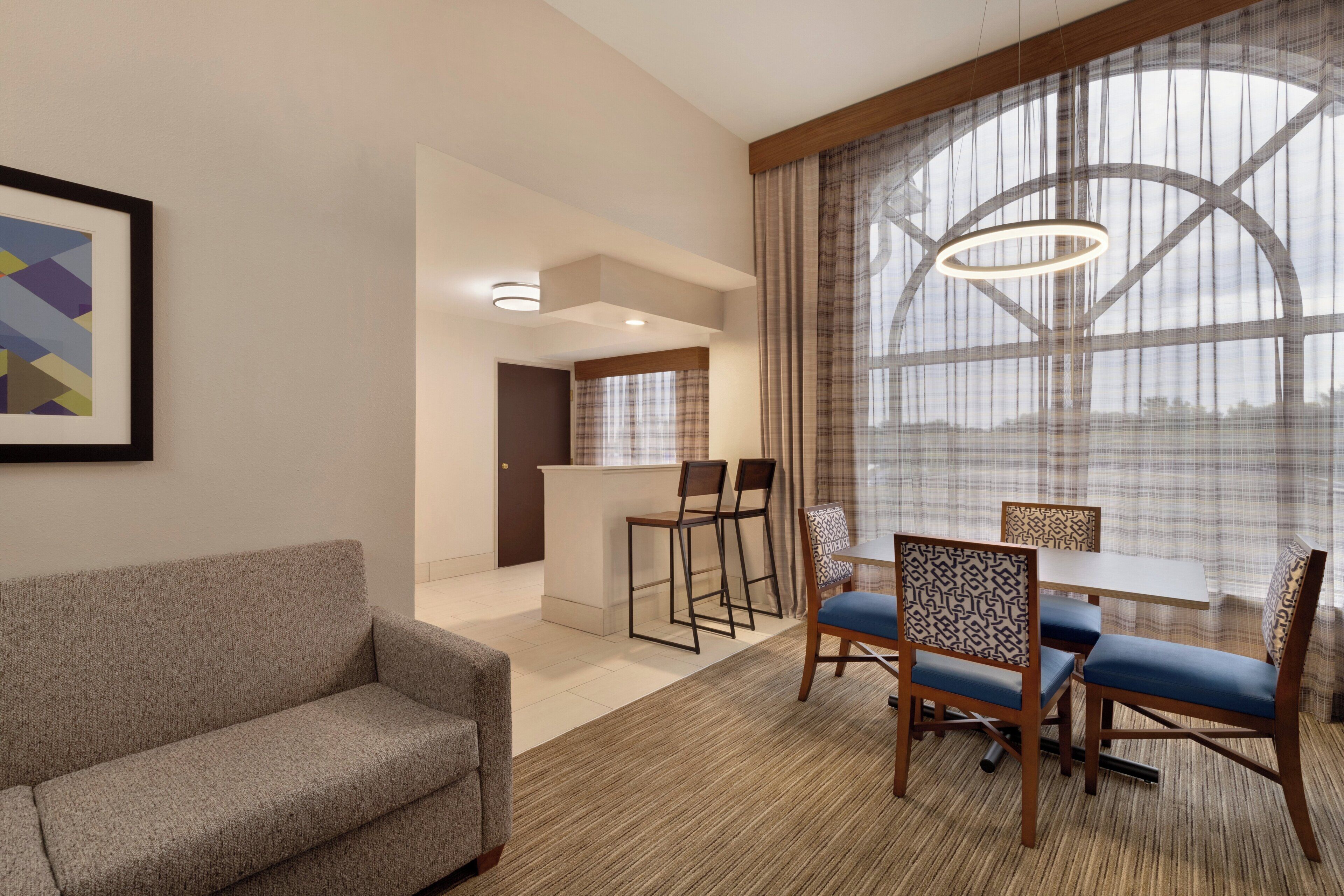 A modern hotel room with a sofa, dining table with chairs, a small kitchen area, and large windows with curtains.