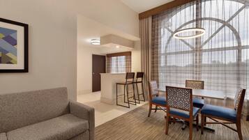 A modern hotel room with a sofa, dining table with chairs, a small kitchen area, and large windows with curtains.
