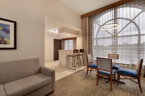 Una habitación de hotel moderna con sofá, mesa de comedor y sillas, una pequeña cocina y ventanas amplias con cortinas.