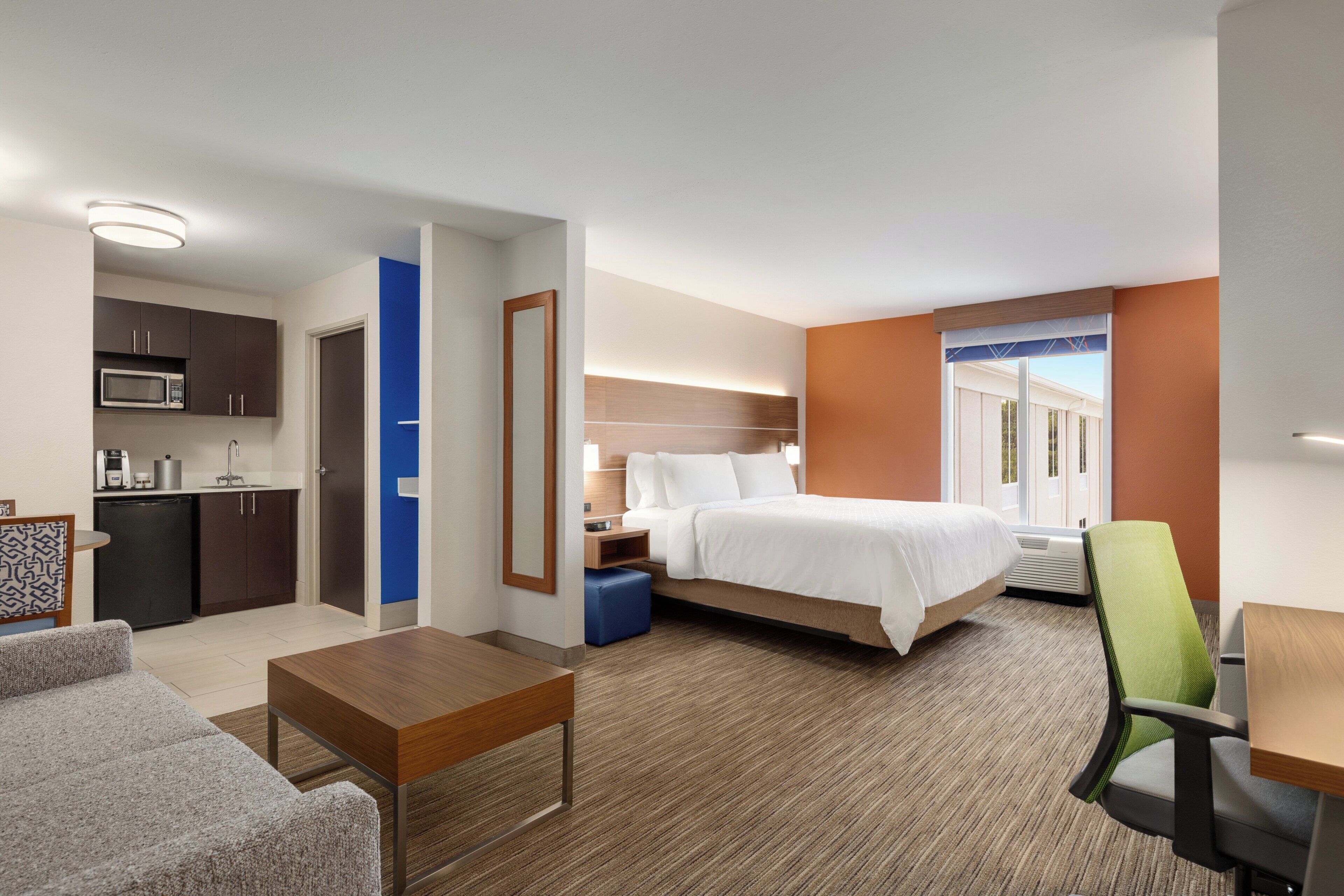 A hotel room with a bed, a desk, a chair, a kitchenette, and a bathroom.
