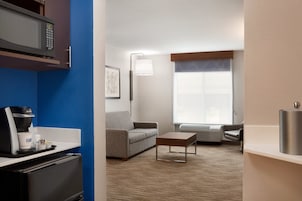 Una habitación de hotel moderna con kitchenette, un sofá gris, una cafetera, un televisor de pantalla plana y un gran ventanal con persianas.