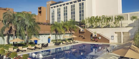 A hotel pool area with lounge chairs, umbrellas, and a large building in the background.