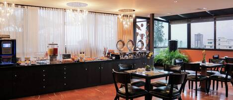 A breakfast bar with a coffee machine, fruit, and pastries. There are dining tables and chairs in the room.