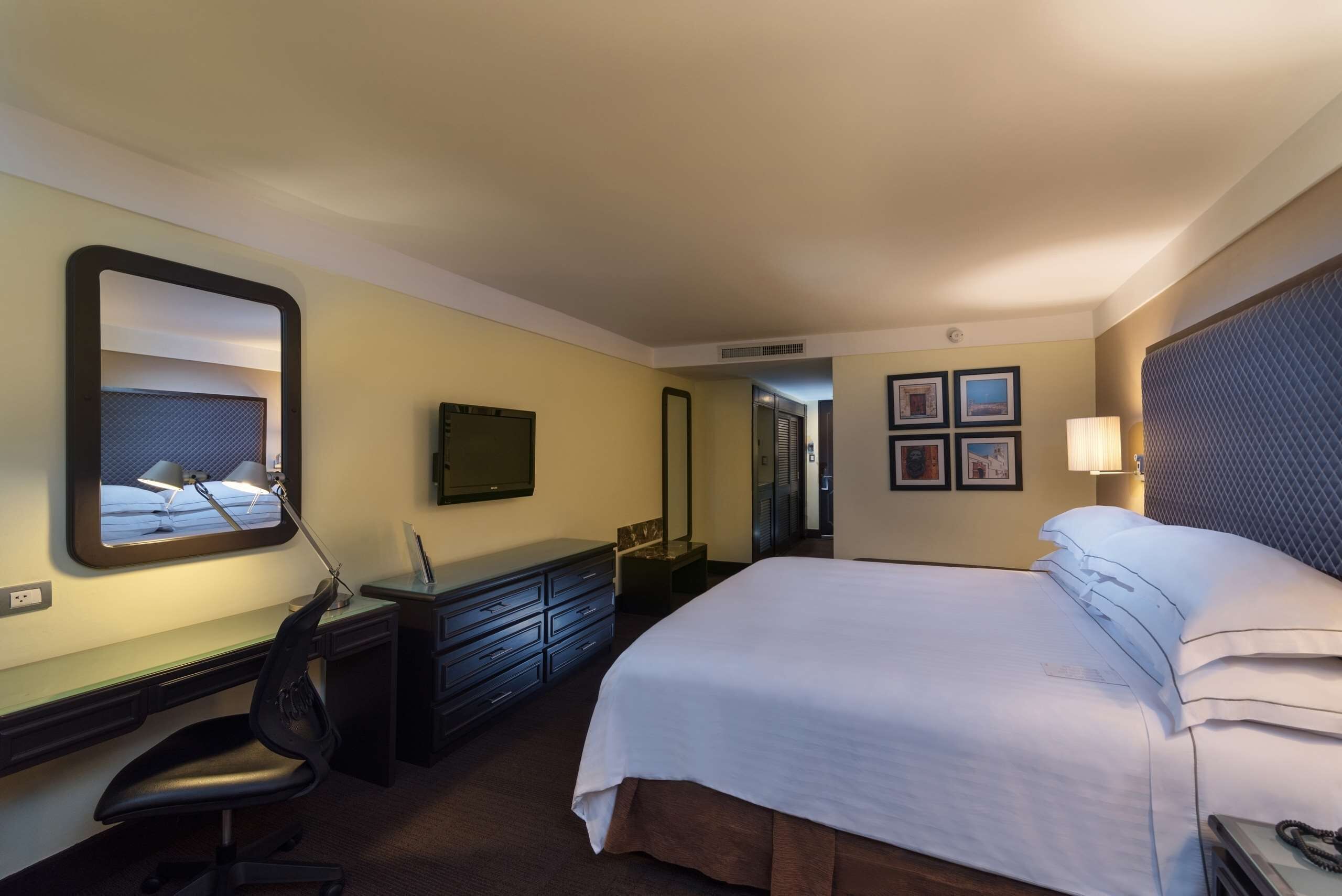 Habitación de hotel con cama, escritorio, silla, cómoda, televisor y cuadros en la pared.