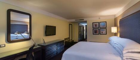 A hotel room with a bed, desk, chair, dresser, TV, and framed pictures on the wall.