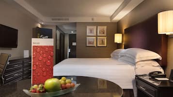 A hotel room with a bed, bedside table with a phone, a bowl of fruit, a red textured column, and framed pictures on the wall.