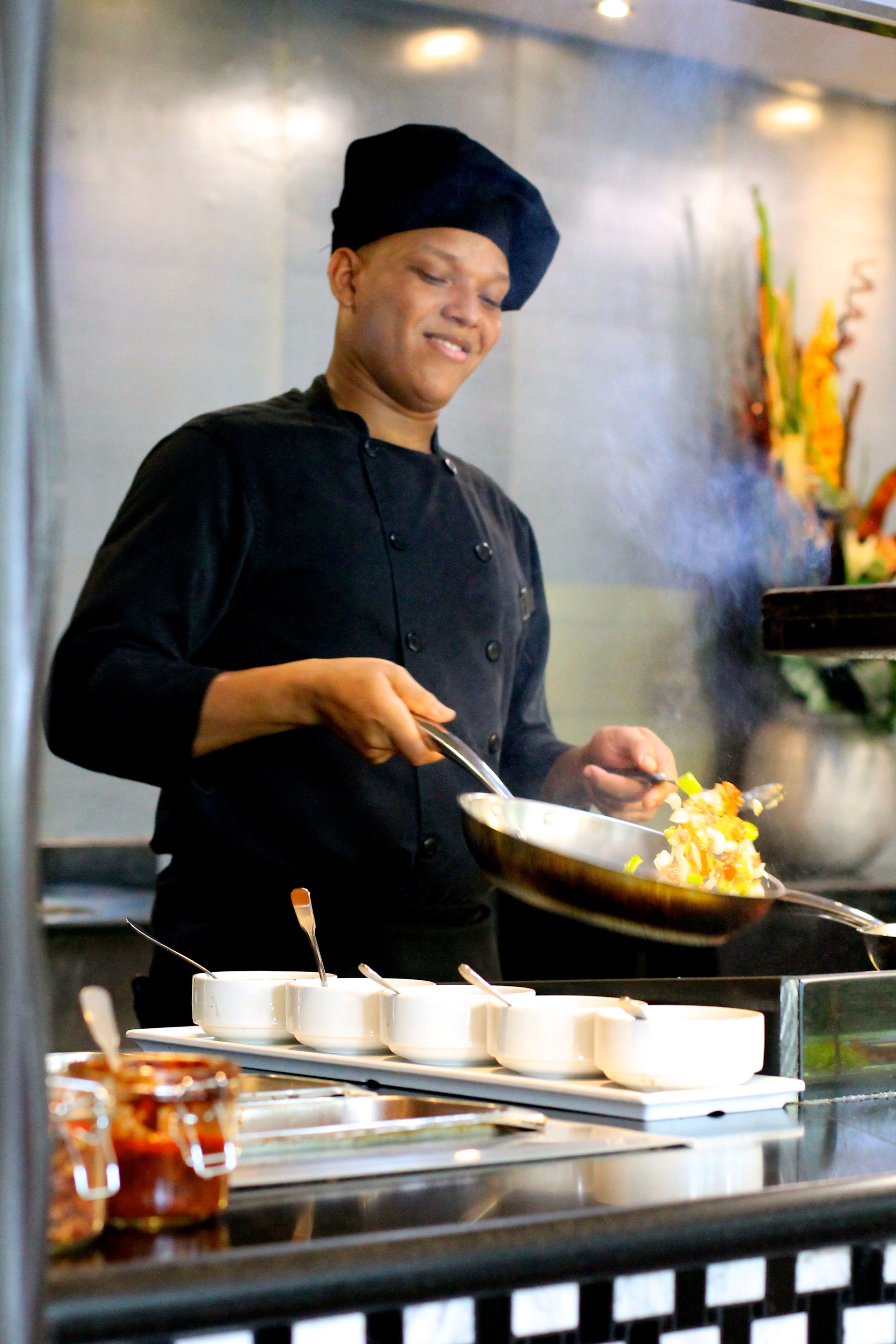 A chef in a black uniform and hat cooking in a professional kitchen with a stainless steel counter, white bowls, and a pot emitting steam.