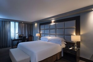 Quarto de hotel com uma cama grande, uma escrivaninha com cadeira e uma mesa de cabeceira com abajur.