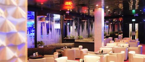 A modern lounge area with white sofas and round tables, featuring a geometric wall design and ambient lighting.