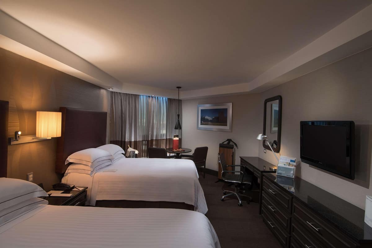 A hotel room with two beds, a desk with a chair, a television, a lamp, and a painting on the wall.