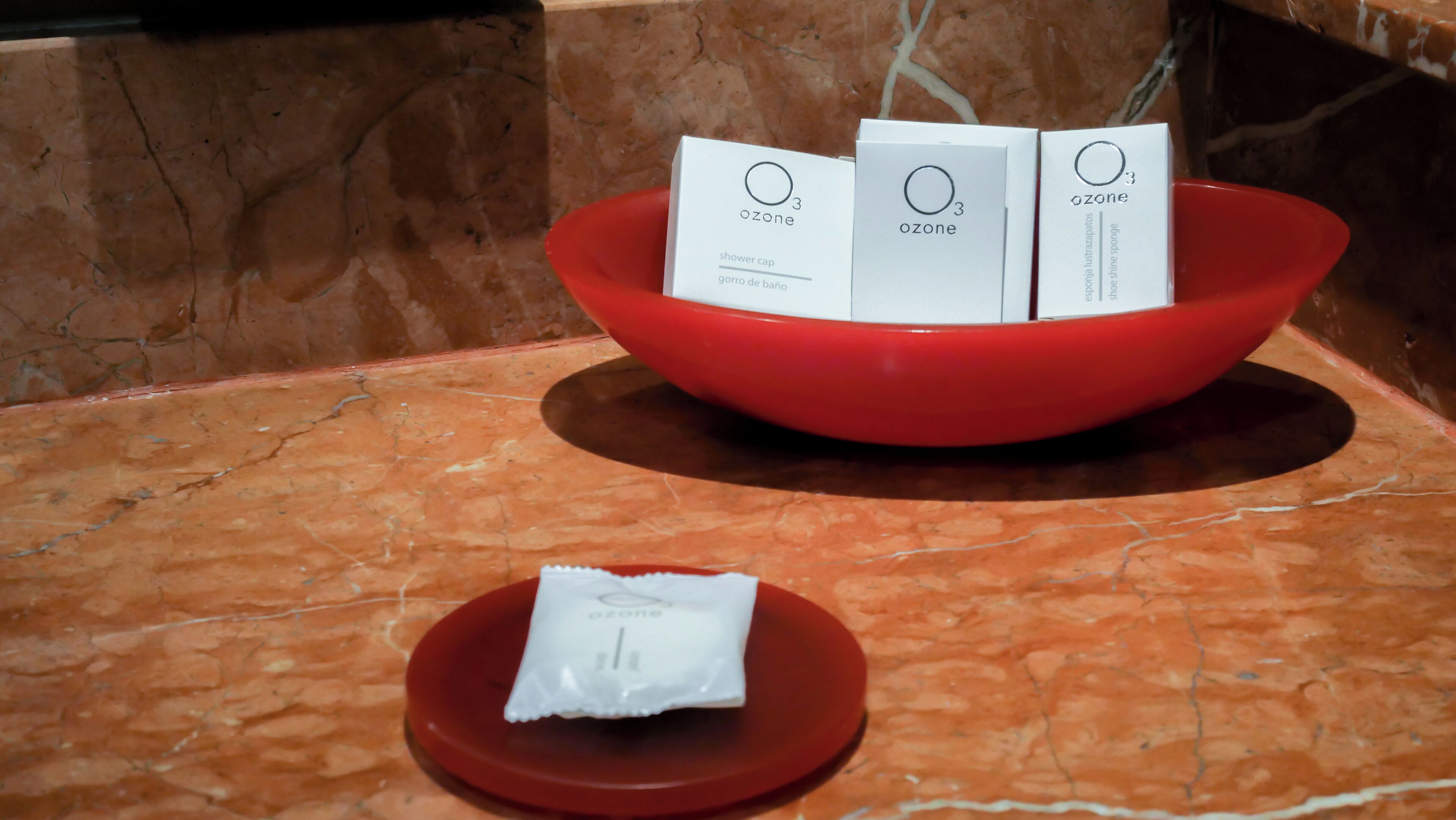 Un tazón rojo que contiene tres cajas blancas etiquetadas 'ozone' con texto en varios idiomas, y un paquete blanco más pequeño sobre un plato rojo a su lado.