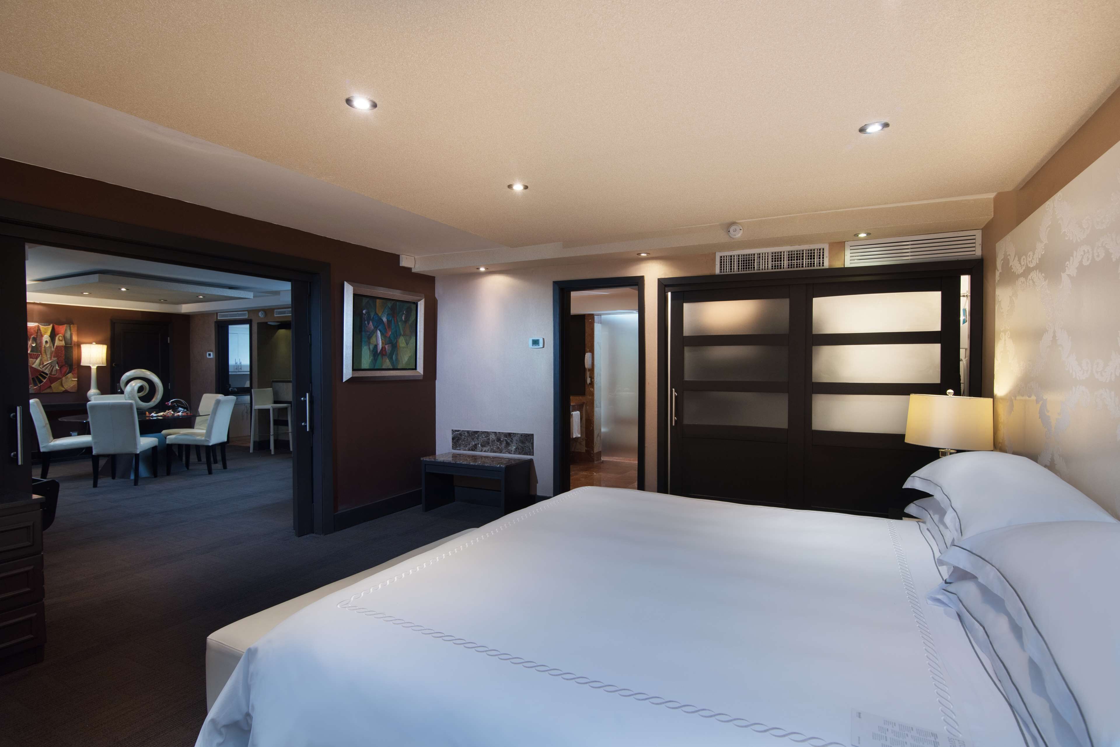 A hotel room with a large bed, a desk with a chair, a dining area with chairs, and a painting on the wall.