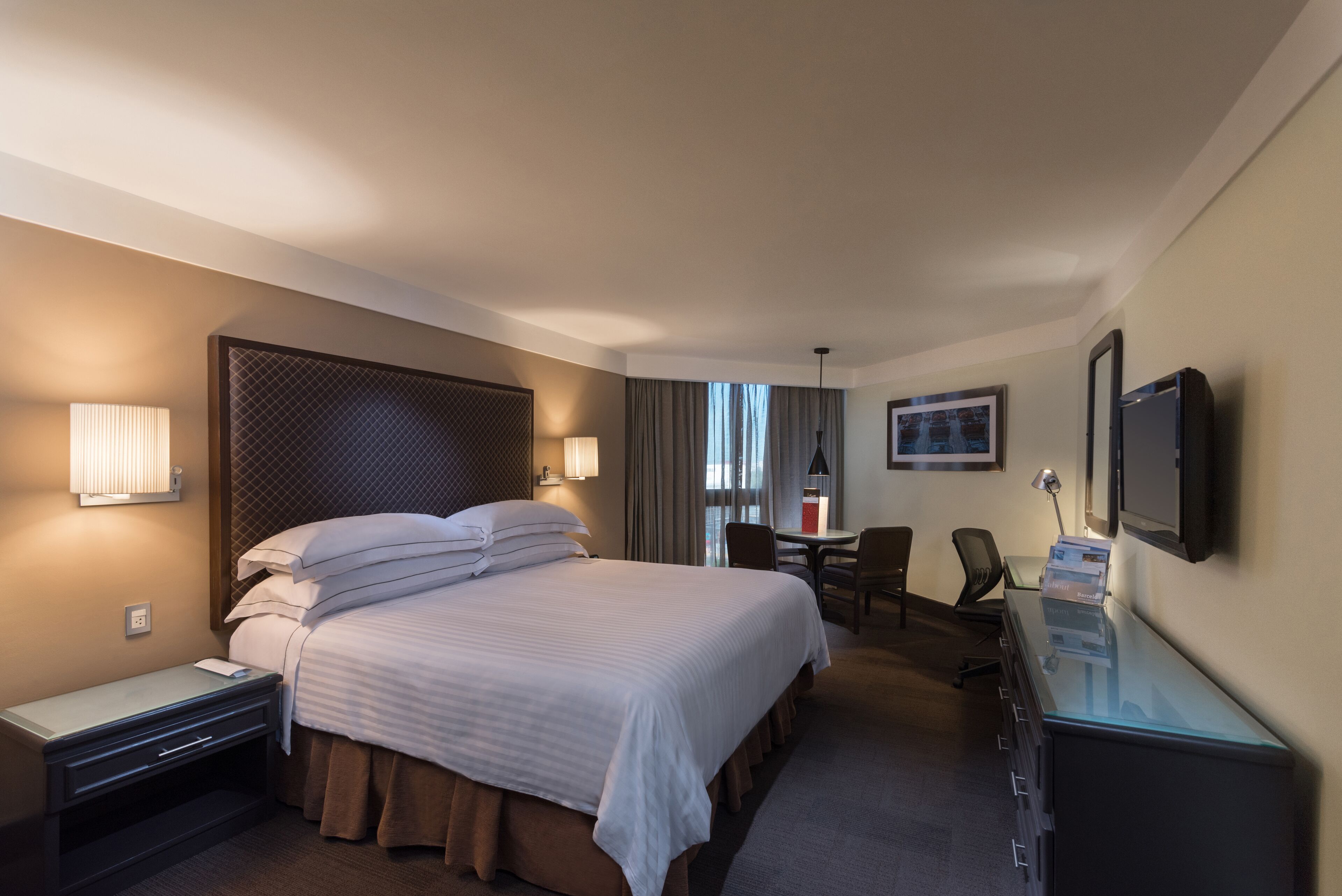 Ett hotellrum med en stor säng, sängbord, ett skrivbord med en dator, en stol och en tv som är monterad på väggen.