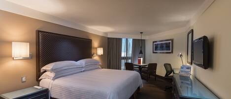 A hotel room with a large bed, bedside tables, a desk with a computer, a chair, and a TV mounted on the wall.