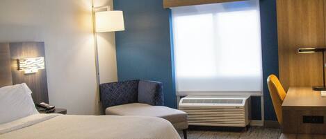 A hotel room with a bed, a desk, a chair, a lamp, a window, and an air conditioning unit.