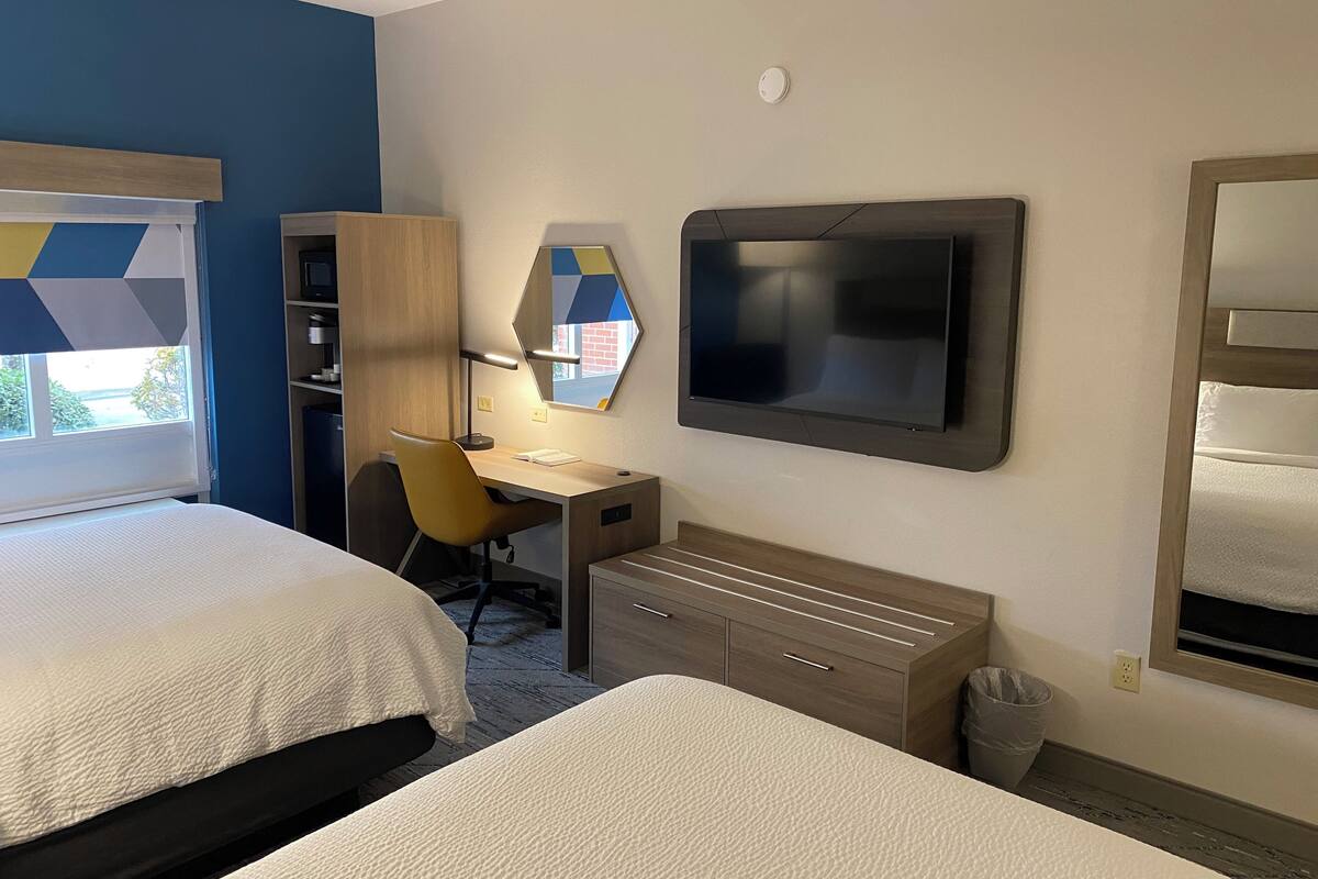 A hotel room with two beds, a desk with a chair, a TV mounted on the wall, and a window with a view of the outside.