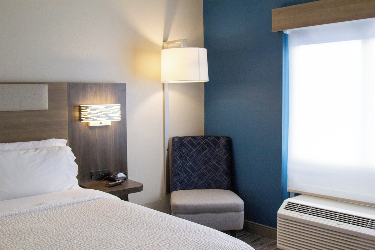 Une chambre d’hôtel avec un lit, une table de chevet, une lampe, une chaise et une fenêtre avec des stores.