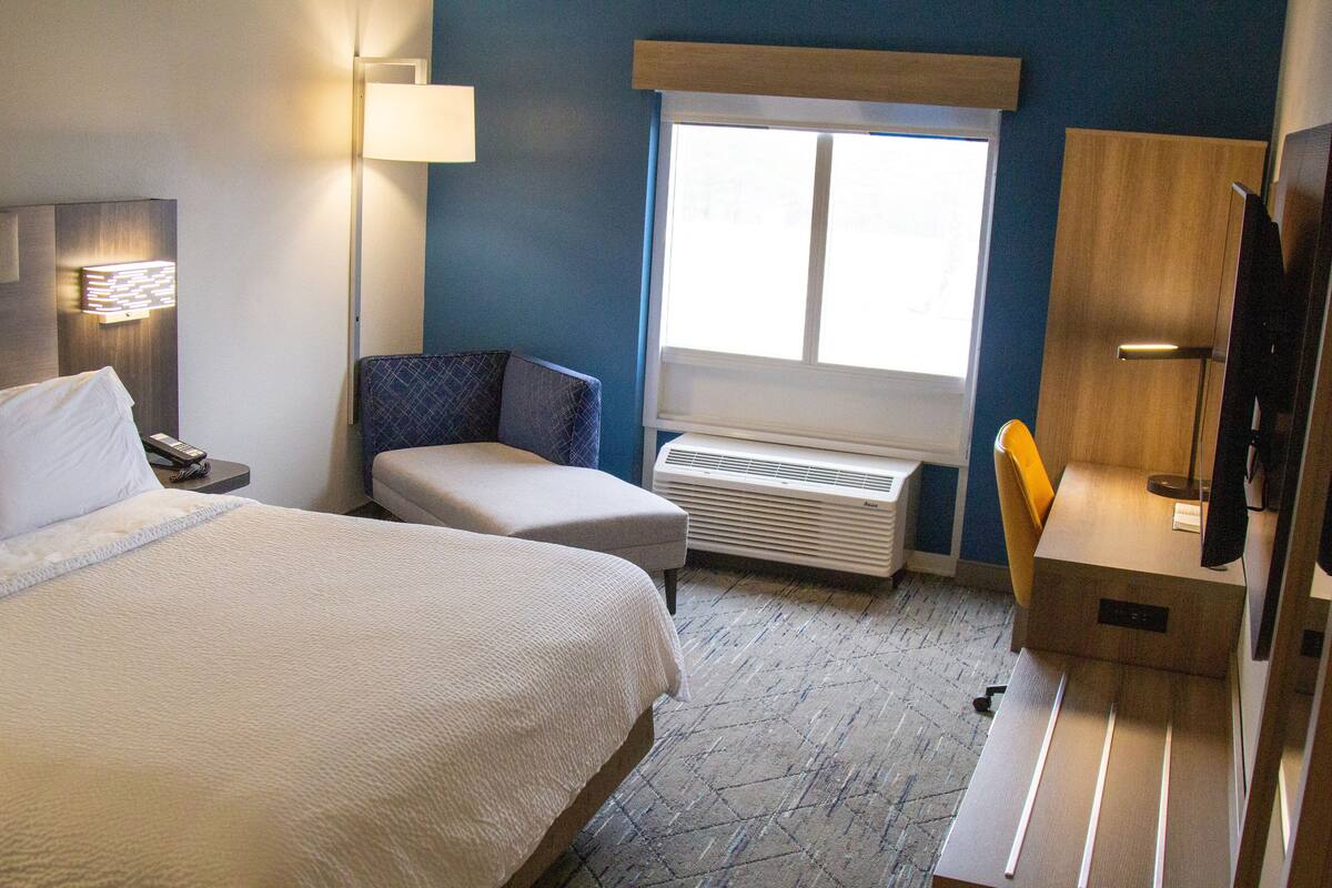 A hotel room with a bed, a desk, a chair, a lamp, and a window.