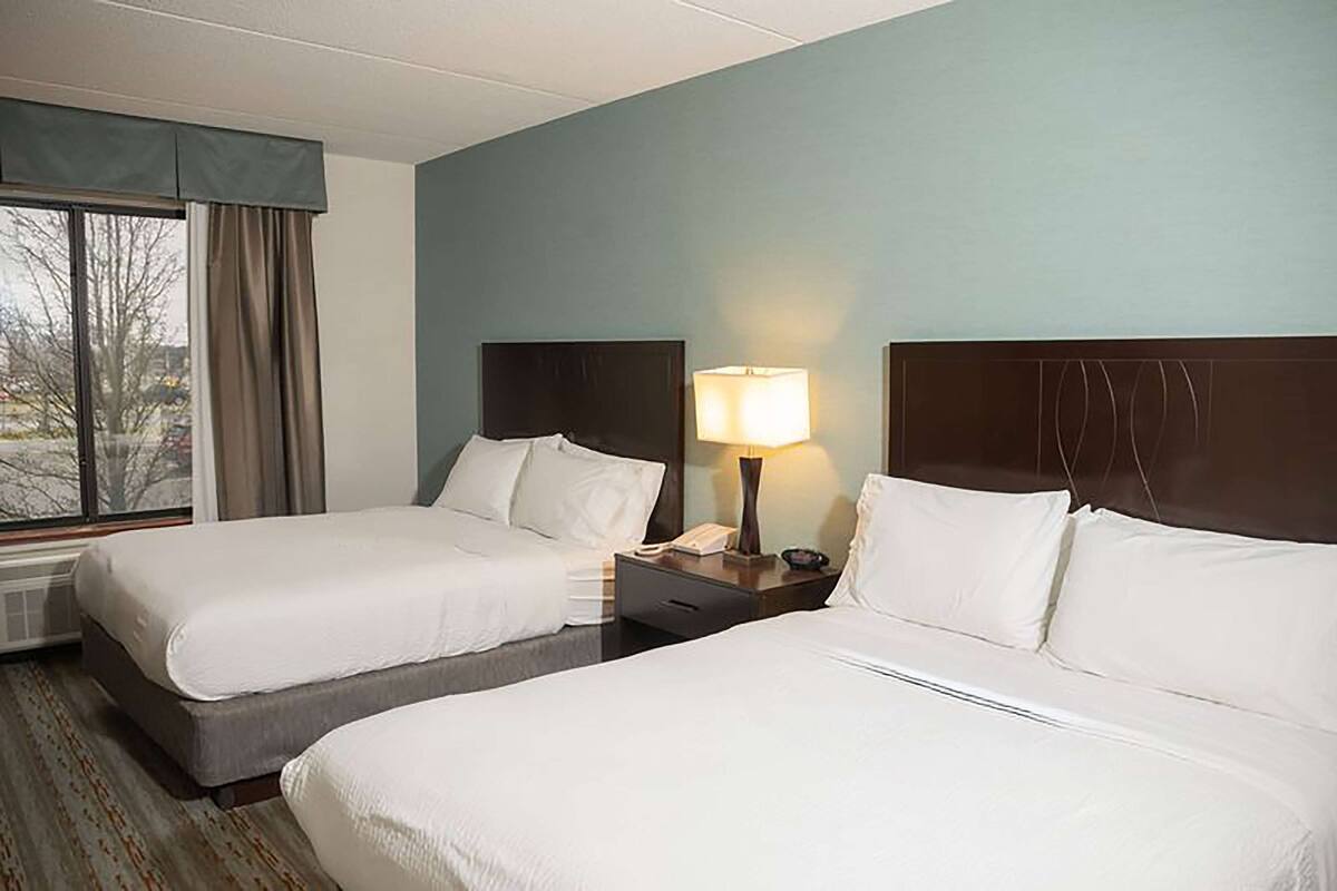 A hotel room with two beds, a window with curtains, a nightstand with a lamp, and a view of the outside.