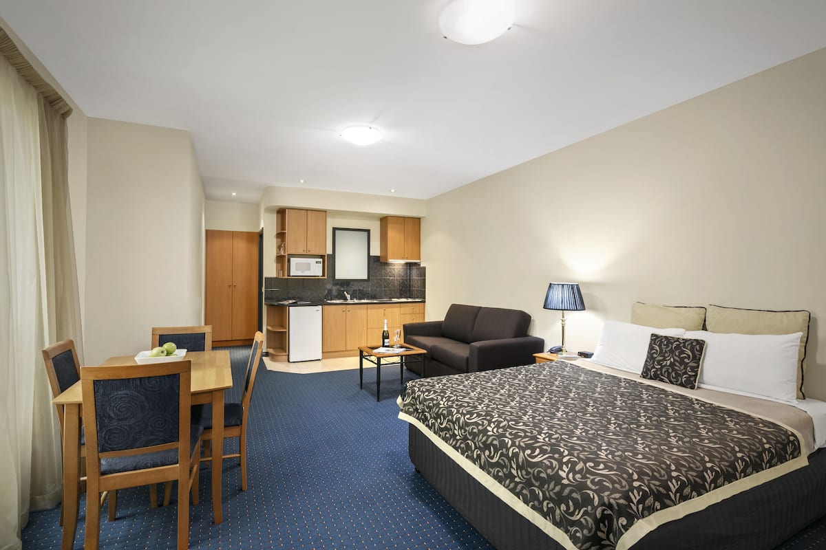 Una habitación de hotel con cama, escritorio, silla, mesita, sofá, lámpara y kitchenette.