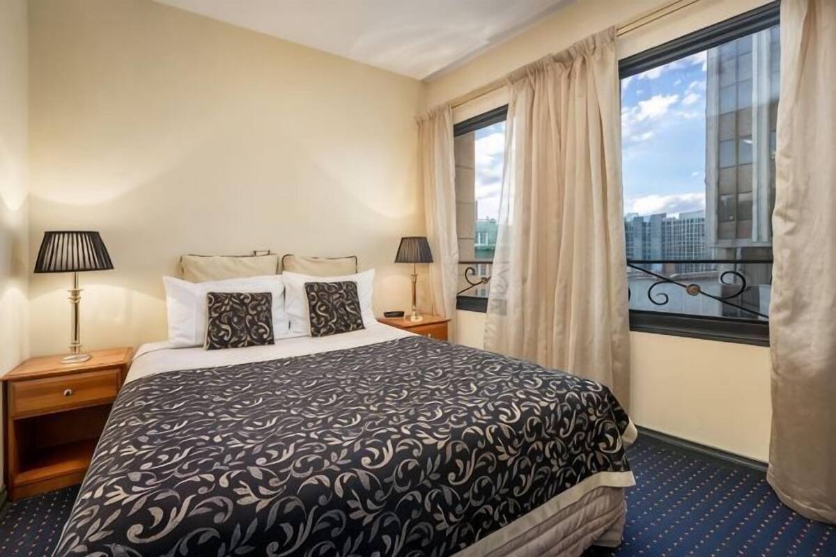 Una habitación de hotel con una cama, dos mesitas de noche, una ventana con cortinas y vistas a la ciudad.