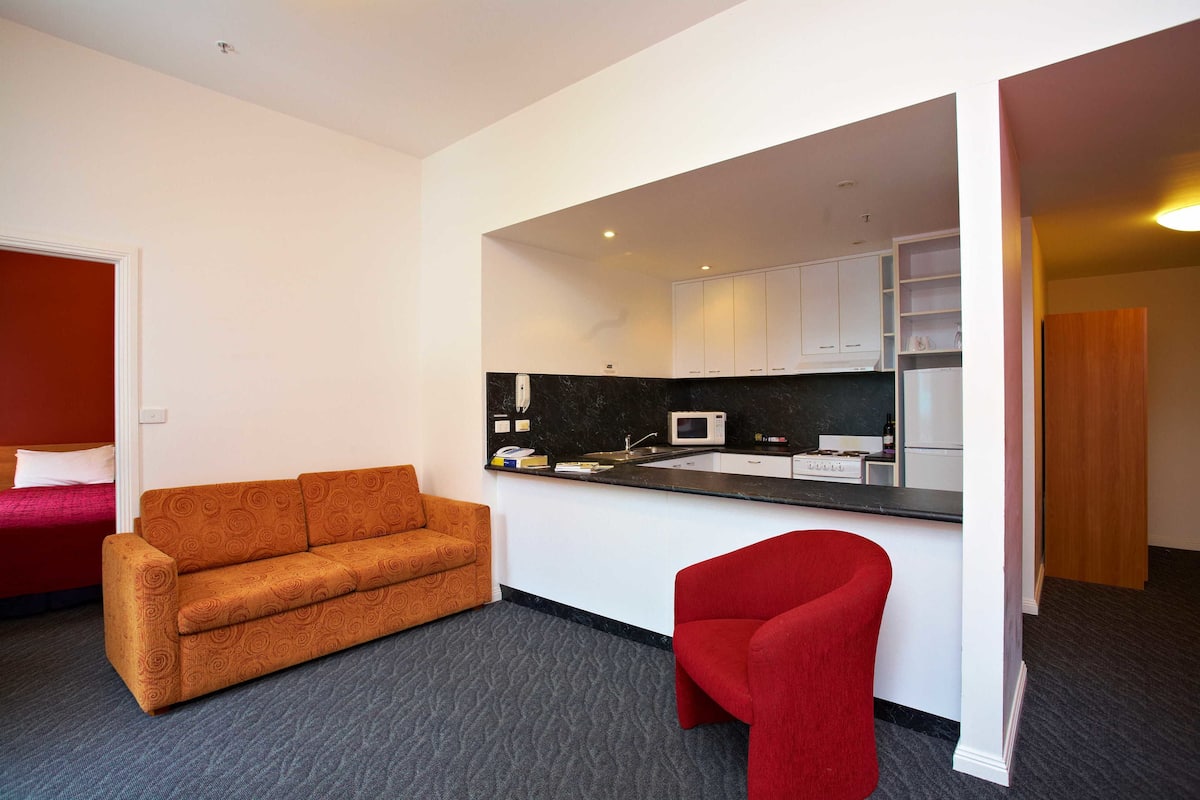 Una habitación compacta con kitchenette, una silla roja, un sofá y una cama con ropa de cama roja.