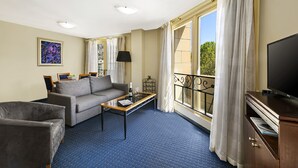 Une chambre d’hôtel équipée d’un canapé, d’un fauteuil, d’une table basse, d’une télévision et d’un balcon avec des rideaux.