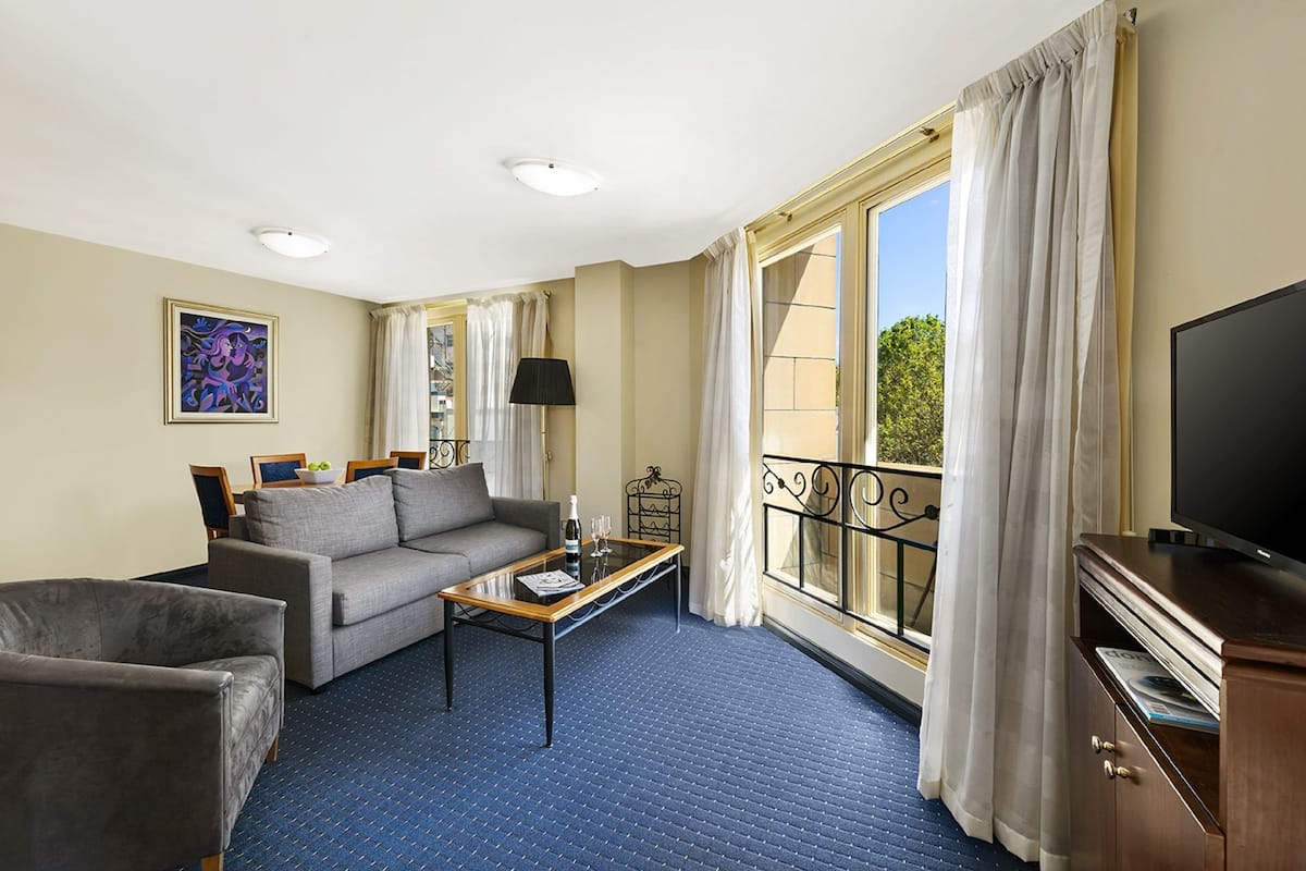 Habitación de hotel con sofá, sillón, mesa de centro, televisión y balcón con cortinas.