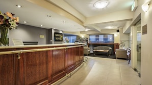 Un espace d’accueil avec un comptoir de réception en bois, un coin salon avec un canapé et des fauteuils, et une vue sur l’extérieur grâce à de grandes fenêtres.