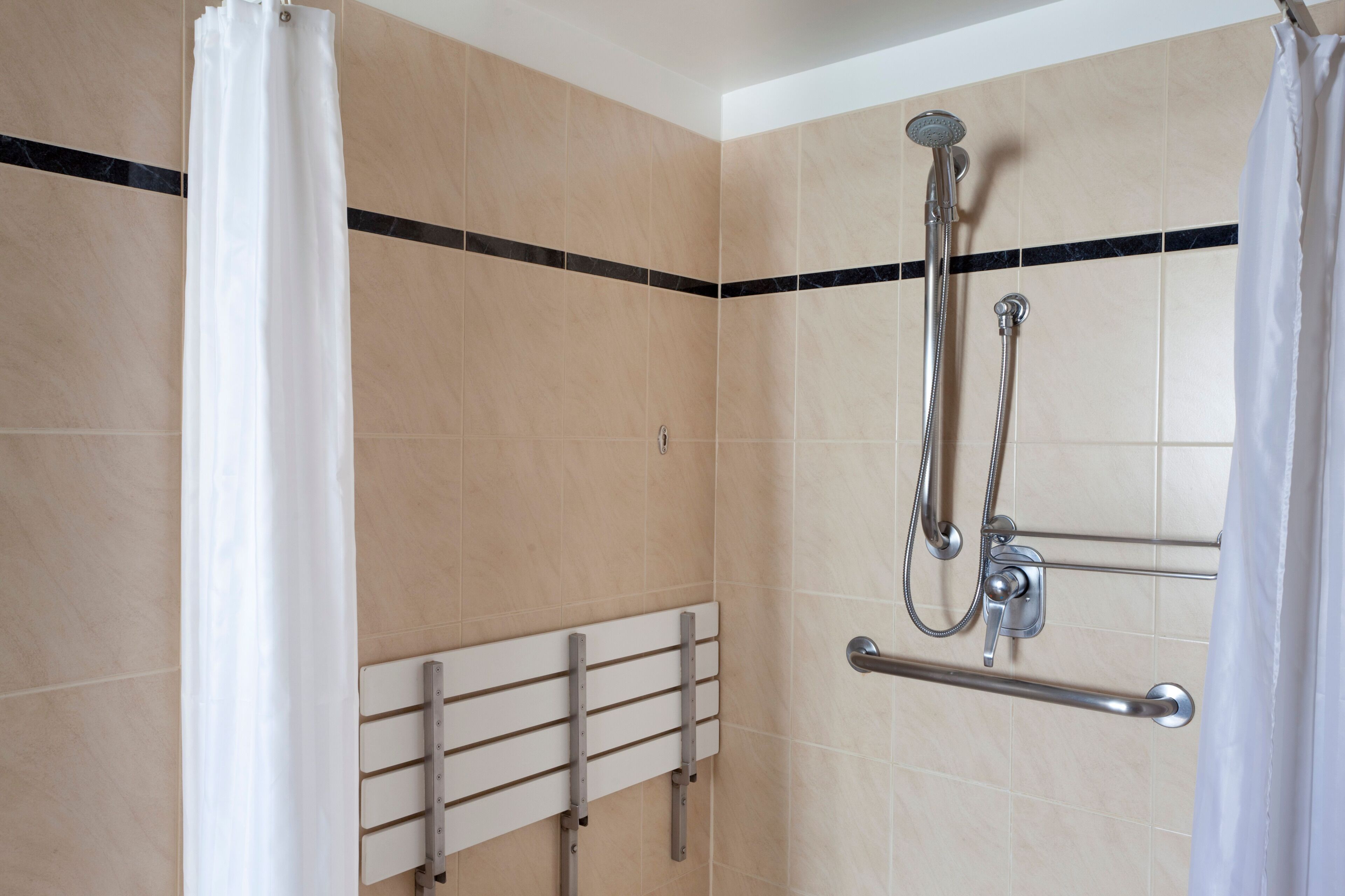 A shower area with beige tiles, a white shower curtain, a handheld showerhead, and a grab bar.