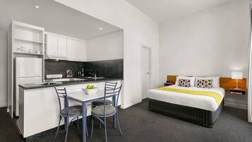 A compact hotel room with a kitchenette, a bed with a yellow throw, and a small table with chairs.